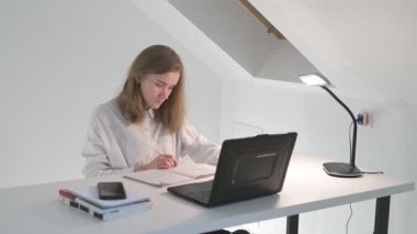 Beyaz tişörtlü liseli kız elinde not defterleri, ders kitapları, akıllı telefon ve dizüstü bilgisayarla masada oturuyor, ödev yapıyor, internetten öğrenim görüyor.