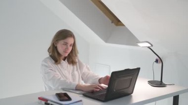 Beyaz tişörtlü liseli kız elinde not defterleri, ders kitapları, akıllı telefon ve dizüstü bilgisayarla masada oturuyor, ödev yapıyor, internetten öğrenim görüyor.