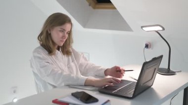 Beyaz tişörtlü liseli kız elinde not defterleri, ders kitapları, akıllı telefon ve dizüstü bilgisayarla masada oturuyor, ödev yapıyor, internetten öğrenim görüyor.