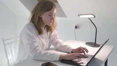 Beyaz tişörtlü liseli kız elinde not defterleri, ders kitapları, akıllı telefon ve dizüstü bilgisayarla masada oturuyor, ödev yapıyor, internetten öğrenim görüyor.