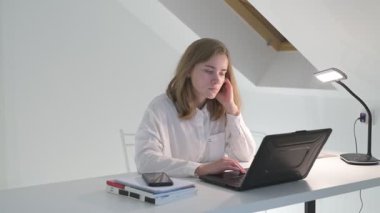 Beyaz tişörtlü liseli kız elinde not defterleri, ders kitapları, akıllı telefon ve dizüstü bilgisayarla masada oturuyor, ödev yapıyor, internetten öğrenim görüyor.