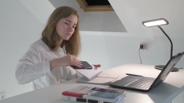 Beyaz tişörtlü liseli kız elinde not defterleri, ders kitapları, akıllı telefon ve dizüstü bilgisayarla masada oturuyor, ödev yapıyor, internetten öğrenim görüyor.