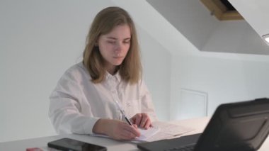 Beyaz tişörtlü liseli kız elinde not defterleri, ders kitapları, akıllı telefon ve dizüstü bilgisayarla masada oturuyor, ödev yapıyor, internetten öğrenim görüyor.