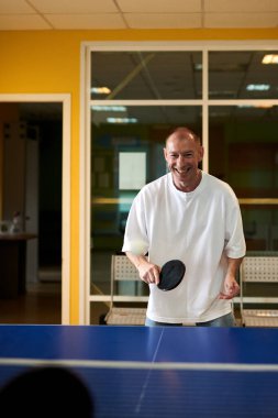 Beyaz tişörtlü bir adam masa tenisi oynamaktan hoşlanır. Pinpon kulübünde dostça bir rekabet.