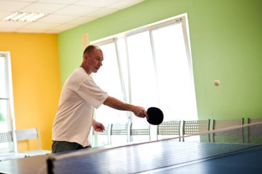 Beyaz tişörtlü bir adam masa tenisi oynamaktan hoşlanır. Pinpon kulübünde dostça bir rekabet.