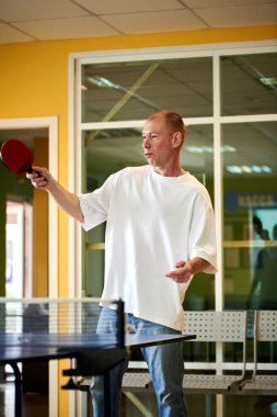 Beyaz tişörtlü bir adam masa tenisi oynamaktan hoşlanır. Pinpon kulübünde dostça bir rekabet.