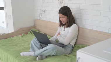 Evde eğitim gören liseli kız. Kız dizüstü bilgisayarla yatakta oturuyor, online kurslar, uzaktan öğrenme.