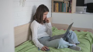 Evde eğitim gören liseli kız. Kız dizüstü bilgisayarla yatakta oturuyor, online kurslar, uzaktan öğrenme.