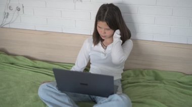 Evde eğitim gören liseli kız. Kız dizüstü bilgisayarla yatakta oturuyor, online kurslar, uzaktan öğrenme.