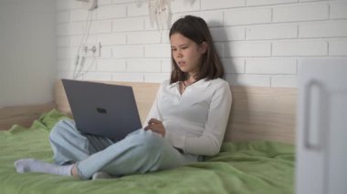Evde eğitim gören liseli kız. Kız dizüstü bilgisayarla yatakta oturuyor, online kurslar, uzaktan öğrenme.