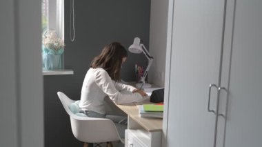 Evde eğitim gören liseli kız. Kız ders kitaplarıyla masada oturup ödev yapıyor.