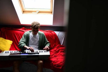Genç adam synthesizer çalıyor, müzisyen evdeki kanepede oturan klavyede şarkı besteliyor.