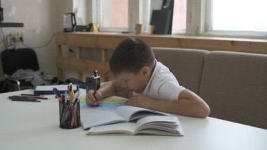 Evde eğitim görmüş bir öğrenci ev ödevini yapar. Çocuk ders kitapları ve renkli kalemlerle masada oturuyor.