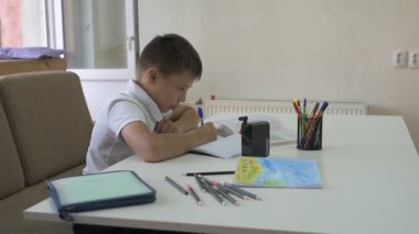 Evde eğitim görmüş bir öğrenci ev ödevini yapar. Çocuk ders kitapları ve renkli kalemlerle masada oturuyor.