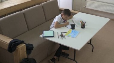 Evde eğitim görmüş bir öğrenci ev ödevini yapar. Çocuk ders kitapları ve renkli kalemlerle masada oturuyor.