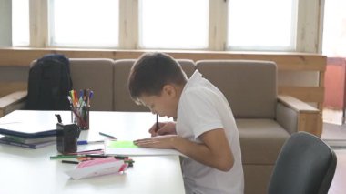 Evde eğitim görmüş bir öğrenci ev ödevini yapar. Çocuk ders kitapları ve renkli kalemlerle masada oturuyor.