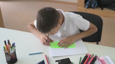 Evde eğitim görmüş bir öğrenci ev ödevini yapar. Çocuk ders kitapları ve renkli kalemlerle masada oturuyor.
