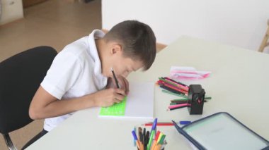 Evde eğitim görmüş bir öğrenci ev ödevini yapar. Çocuk ders kitapları ve renkli kalemlerle masada oturuyor.