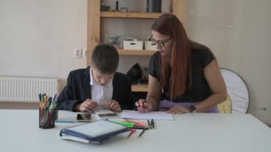 Evde eğitim görmüş bir çocuk ödevini masada annesiyle yapıyor. Çocuk evde bir öğretmenle ders çalışıyor.