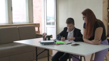 Evde eğitim görmüş bir çocuk ödevini masada annesiyle yapıyor. Çocuk evde bir öğretmenle ders çalışıyor.