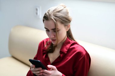 Evdeki kanepede elinde cep telefonuyla oturan genç bir kadın.