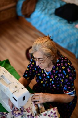 Emeklilik yaşında yaşlı bir kadın elektrikli dikiş makinesine elbise dikiyor.