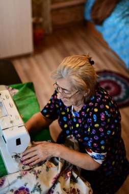 Emeklilik yaşında yaşlı bir kadın elektrikli dikiş makinesine elbise dikiyor.