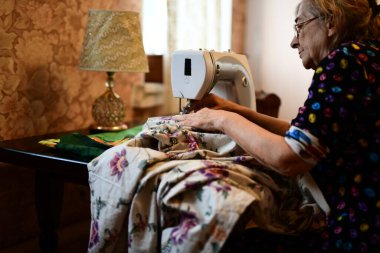 Emeklilik yaşında yaşlı bir kadın elektrikli dikiş makinesine elbise dikiyor.