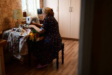 Emeklilik yaşında yaşlı bir kadın elektrikli dikiş makinesine elbise dikiyor.