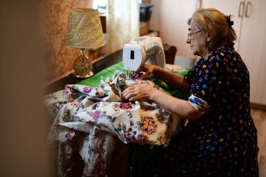 Emeklilik yaşında yaşlı bir kadın elektrikli dikiş makinesine elbise dikiyor.