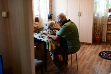 Emeklilik yaşında yaşlı bir adam elektrikli dikiş makinesine elbise dikiyor.