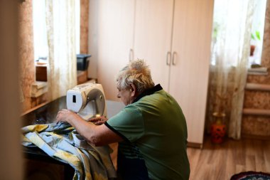 Emeklilik yaşında yaşlı bir adam elektrikli dikiş makinesine elbise dikiyor.