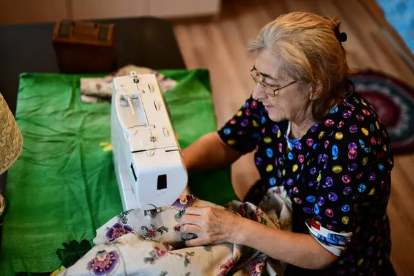 Emeklilik yaşında yaşlı bir kadın elektrikli dikiş makinesine elbise dikiyor.