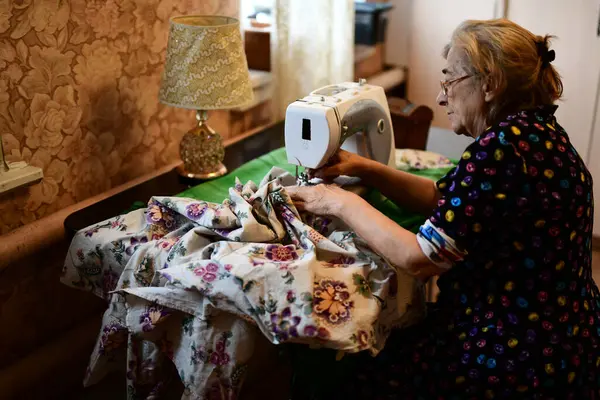 Emeklilik yaşında yaşlı bir kadın elektrikli dikiş makinesine elbise dikiyor.