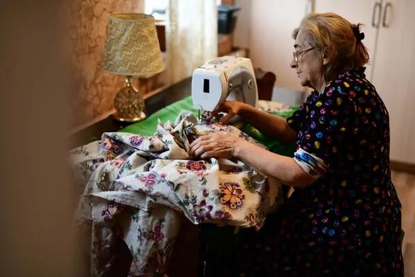 Emeklilik yaşında yaşlı bir kadın elektrikli dikiş makinesine elbise dikiyor.