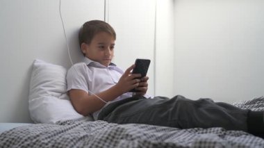 Küçük okul çocuğu evde yatarken cep telefonu ekranını seyretmeye dalmış.