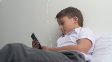 Küçük okul çocuğu evde yatarken cep telefonu ekranını seyretmeye dalmış.