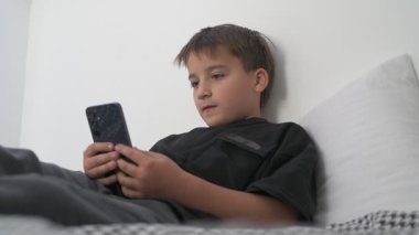 Küçük okul çocuğu evde yatarken cep telefonu ekranını seyretmeye dalmış.