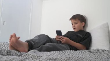 Küçük okul çocuğu evde yatarken cep telefonu ekranını seyretmeye dalmış.