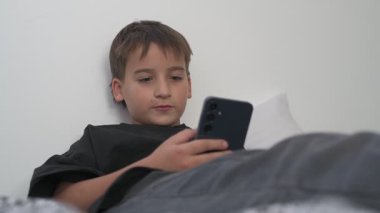 Küçük okul çocuğu evde yatarken cep telefonu ekranını seyretmeye dalmış.