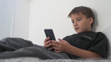 Küçük okul çocuğu evde yatarken cep telefonu ekranını seyretmeye dalmış.