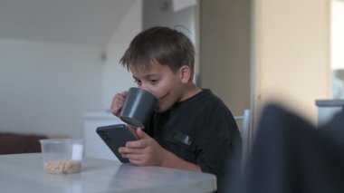 Okul çocuğu okuldan önce mutfakta kahvaltı hazırlıyor, kahve yapıyor ve cep telefonunu kontrol ediyor.