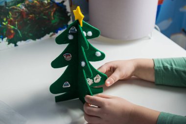 Çocuk, çocuk odasında el yapımı Noel ağacını kendi elleriyle tutuyor. Kutlama kartı için Xmas oyuncakları dekorasyonu yapıyorum. Mutlu Noeller ve mutlu yıllar. El yapımı konsept.