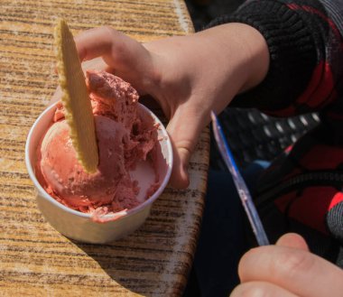 Küçük çocuk masanın üzerinde kaputta pembe dondurma yiyor. Pembe ceketli iki el. Bahar güneşinde, kafenin dışında bir dondurma..