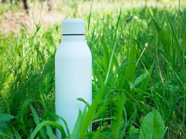 Beyaz termodinamik paslanmaz şişe yeşil çimenlikte ferahlatıcı su ile arka planda.