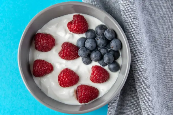 Doğal taze Yunan yoğurdu, ahududulu, böğürtlenli, koyu tabakta ve sağlıklı kahvaltı için peçeteli. Mavi arkaplan.