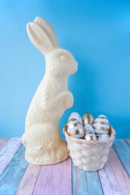 Mavi arka planda Paskalya tavşanı şeklinde beyaz çikolata ve paskalya altını ve beyaz yumurta sepeti..