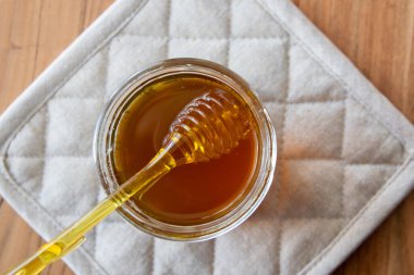 Sarı kaşıklı bal peteği olan bal kavanozu ahşap masada ve pamuk bej peçetede kalsın..