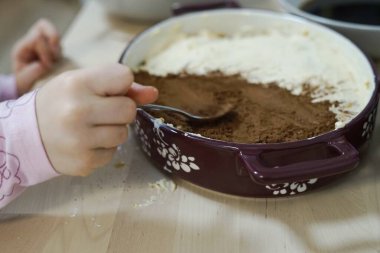 Küçük kız üzerinde kakao tozu olan ev yapımı tiramisu keki yapmaya yardım ediyor ve ahşap masaya dökülüyor. Aile yadigarı ev yapımı çay pişiriyor. Yüksek kalite fotoğraf