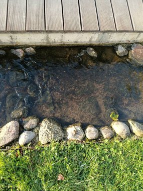 Pürüzsüz kayalar ve yemyeşil otlarla çevrili sakin bir su akıntısı üzerinde zarif bir şekilde uzanan güzel, kırsal ahşap bir köprü.